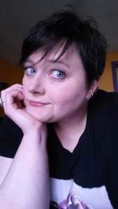 Christin is a middle-aged white woman with short, dark hair. She's wearing a black shirt with a pink Jason Voorhees graphic on it. She's sitting with her chin in her hand giving the camera an "Oh really?" face.